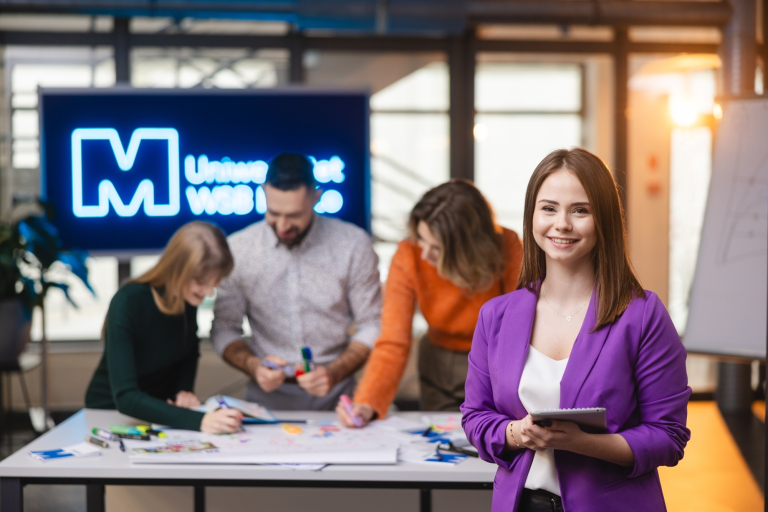 Pośrednictwo pracy | Uniwersytet WSB Merito Opole: Na pierwszym planie uśmiechnięta dziewczyna trzymająca notatnik, w tle grupa studentów pracująca nad projektem