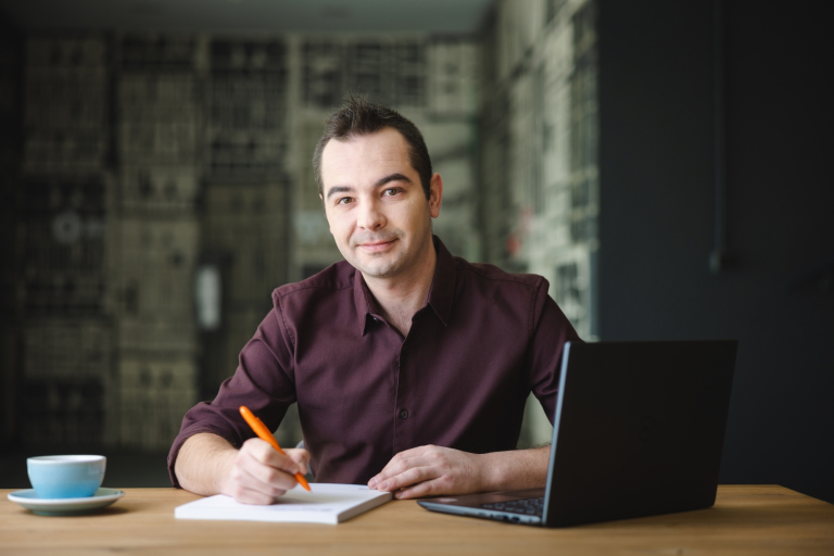 Sztuczna inteligencja w marketingu cyfrowym | Studia podyplomowe w Opolu | Uniwersytet WSB Merito Opole: Mężczyzna zapisuje informacje w notatniku, przed nim stoi otwarty laptop