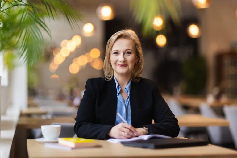 Studia podyplomowe w Opolu | Uniwersytet WSB Merito Opole: Program firma - kobieta siedząca w kawiarni, w dłoni trzyma długopis, przed nią leżą książki i laptop