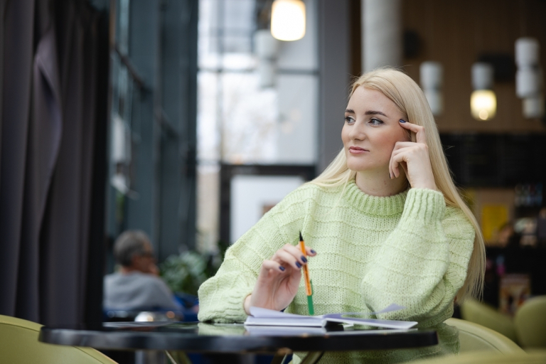 Zamówienia publiczne | Studia podyplomowe w Opolu | Uniwersytet WSB Merito Opole: Kobieta siedząca w kawiarni, ma przed sobą otwarty notatnik, patrzy w dal