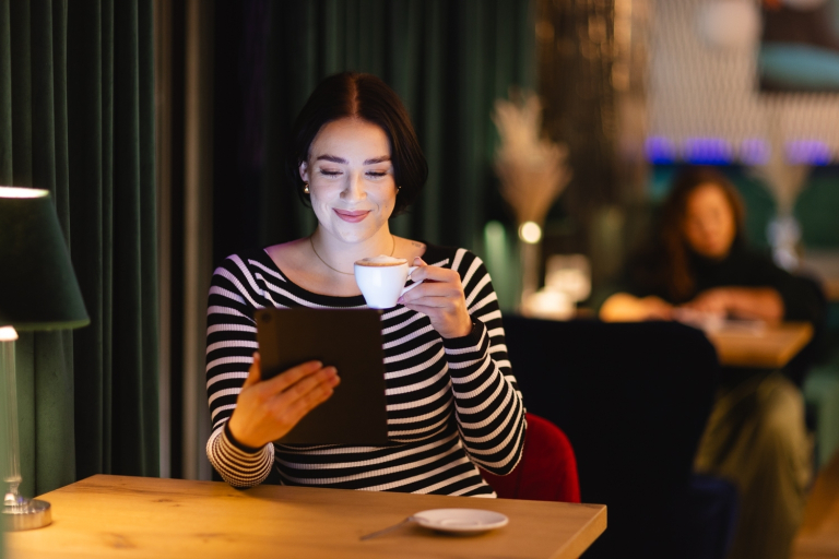 Zarządzanie | Studia II stopnia w Opolu | Uniwersytet WSB Merito Opole: Dziewczyna w kawiarni, w jednej ręce trzyma tablet w drugiej filiżankę kawy