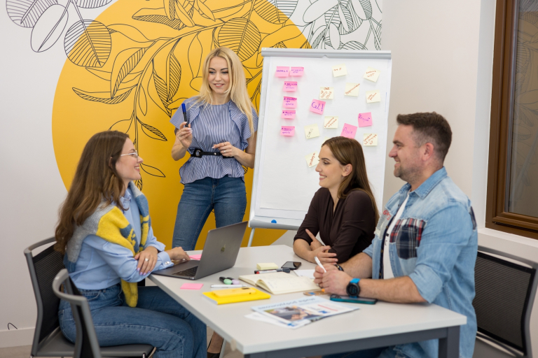 Finanse i rachunkowość | Studia II stopnia ze studiami podyplomowymi w Opolu | Uniwersytet WSB Merito Opole: Grupa osób siedzących przy stole, wymieniają się pomysłami, na stole leżą notatki, jedna z kobiet stoi przy białej tablicy