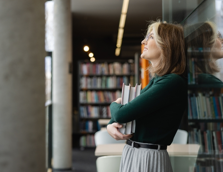 Kompetencje przyszłości - przewaga w świecie zmian | Studia podyplomowe w Opolu | Uniwersytet WSB Merito Opole: Uczestniczka studiów podyplomowych stoi i opera się plecami o półkę w bibliotece, trzymając w ręce książki