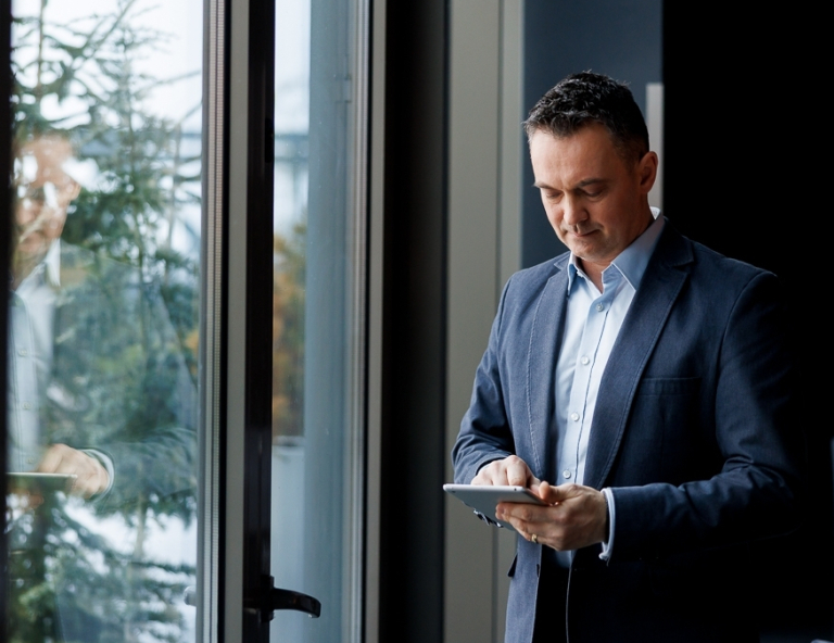 Inżynieria Oprogramowania dla prędkości produktu | Studia podyplomowe w Opolu | Uniwersytet WSB Merito Opole: Uczestnik studiów podyplomowych opiera się o okno i spogląda w tableta