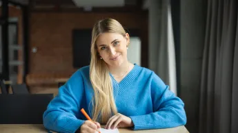 Psychologia negocjacji i mediacji | Studia I stopnia w Poznaniu | Uniwersytet WSB Merito w Poznaniu: kobieta w niebieskim swetrze siedzi przy stole i z uśmiechem sporządza notatki