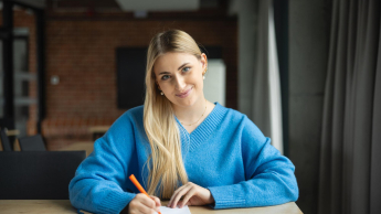 Psychologia negocjacji i mediacji | Studia I stopnia w Poznaniu | Uniwersytet WSB Merito w Poznaniu: kobieta w niebieskim swetrze siedzi przy stole i z uśmiechem sporządza notatki