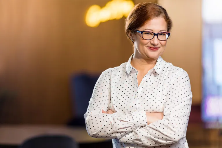 Oferty pracy | Uniwersytet WSB Merito w Poznaniu: Kobieta w okularach uśmiecha się, stojąc z założonymi rękami w nowoczesnym wnętrzu.