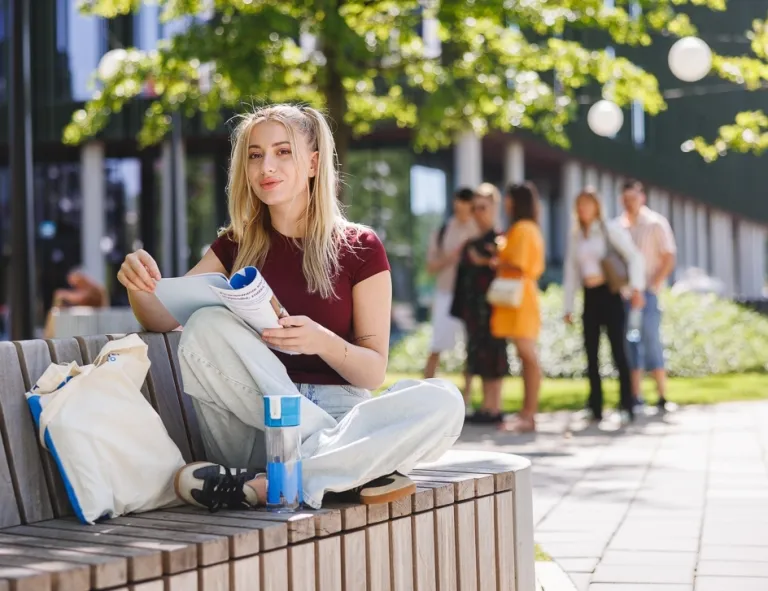 Strona główna | Uniwersytet WSB Merito w Poznaniu: Studentka WSB Merito siedzi na ławce w plenerze i przegląda informator, w tle grupa osób wesoło rozmawia