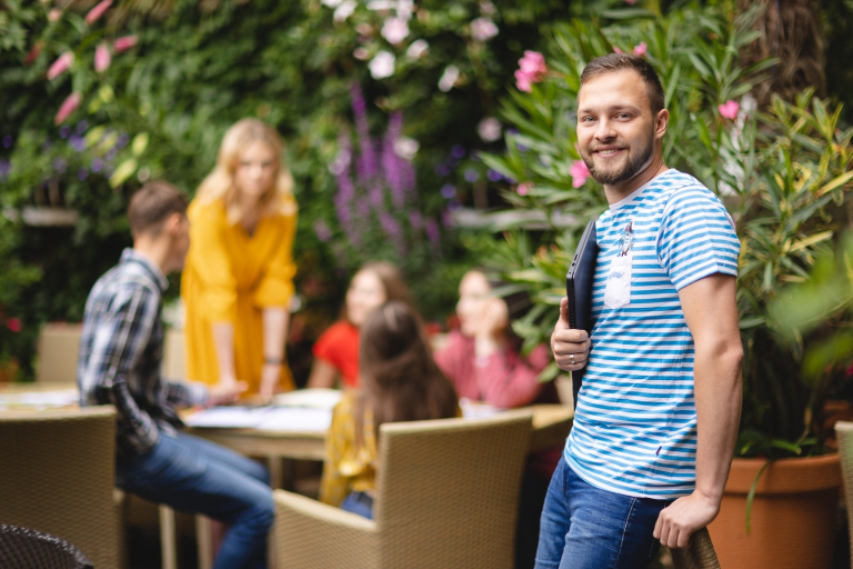 Strefa studenta | Uniwersytet WSB Merito w Poznaniu: Student WSB Merito trzyma komputer w ręce i uśmiecha się, w tle jego znajomi siedzą przy stoliku w kawiarni pełnej roślin