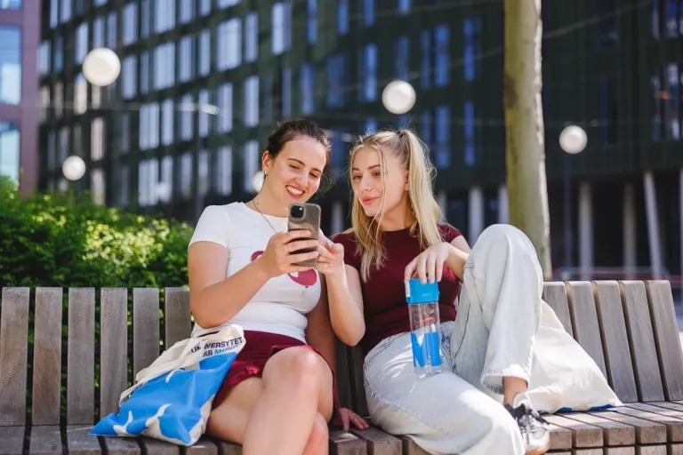 Innowacje i AI w biznesie | Uniwersytet WSB Merito w Poznaniu: Dwie dziewczyny siedzą na ławce, jedna pokazuje drugiej coś w telefonie