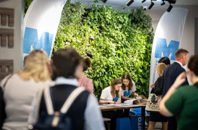 Sustainable and Socially Responsible Development in the Network of Socioeconomic Relations | Uniwersytet WSB Merito: Dwie studentki pochylone przy stole nad materiałami konferencyjnymi wśród przechodzących ludzi