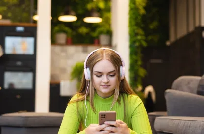 Poradniki - dzielimy się wiedzą | Uniwersytet WSB Merito: Dziewczyna siedząca z telefonem w dłoni, odtwarzająca materiał przez słuchawki