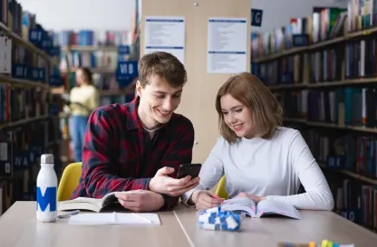 Poradniki - dzielimy się wiedzą | Uniwersytet WSB Merito: Dwie osoby patrzące w telefon, siedząc w bibliotece