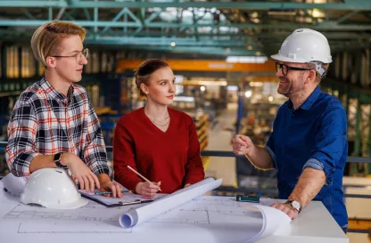 Poradniki - dzielimy się wiedzą | Uniwersytet WSB Merito: Grafika główna: Uniwersytety WSB Merito, praca po studiach, inżynieria produkcji, 2 mężczyzn i kobieta rozmawiają w hali produkcyjnej