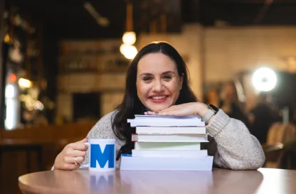 Poradniki - dzielimy się wiedzą | Uniwersytet WSB Merito: Uśmiechnięta kobieta przy stole opierająca się na stosie książek z kubkiem Merito w dłoni