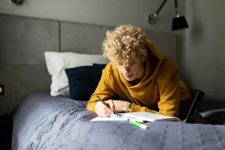 Wypalenie akademickie - jak temu zapobiec? | Blog "okiem praktyków" | Uniwersytet WSB Merito: Młody chłopak leżący na łóżku spoglądający w notatki