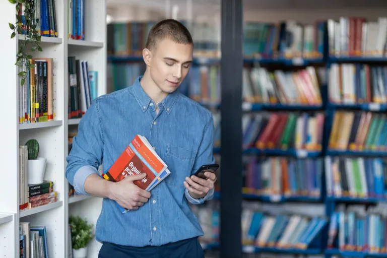 Czemu warto kontynuować studia i wybrać się na magisterkę? | Uniwersytet WSB Merito: Chłopak w bibliotece trzymający telefon