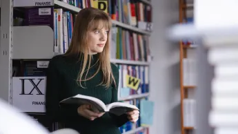 Bibliotekoznawstwo w szkole | Studia podyplomowe w Szczecinie | Uniwersytet WSB Merito Szczecin: Studentka Uniwersytetu WSB Merito spogląda za okno, przeglądając książkę w bibliotece.
