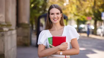 Pedagogika opiekuńczo-wychowawcza i terapia zajęciowa | Studia I stopnia w Szczecinie | Uniwersytet WSB Merito Szczecin: Uśmiechnięta Studentka trzyma książki stojąc w parku.