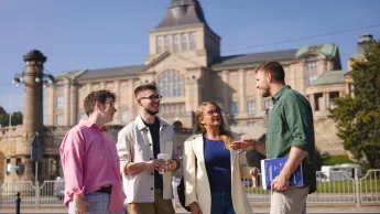 Zarządzanie logistyką w służbach mundurowych | Studia podyplomowe w Szczecinie | Uniwersytet WSB Merito Szczecin: Czworo studentów rozmawia na tle gmachu Wałów Chrobrego w słoneczny dzień.