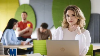 SAP S/4HANA – nowoczesny system ERP | Studia podyplomowe w Szczecinie | Uniwersytet WSB Merito Szczecin: Studentka pracuje przy laptopie w nowoczesnej przestrzeni z innymi osobami w tle.