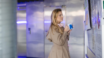 Studentka analizuje informacje na tablicy, trzymając kubek i słuchawki, w nowoczesnym wnętrzu uczelni.