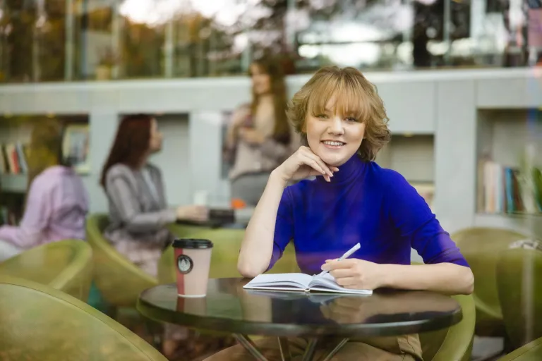 Edukacja i rehabilitacja osób z niepełnosprawnością intelektualną – dla pedagogów specjalnych | Studia podyplomowe w Szczecinie | Uniwersytet WSB Merito Szczecin: Studentka Uniwersytetu WSB Merito notuje w zeszycie pijąc kawę przy stoliku.