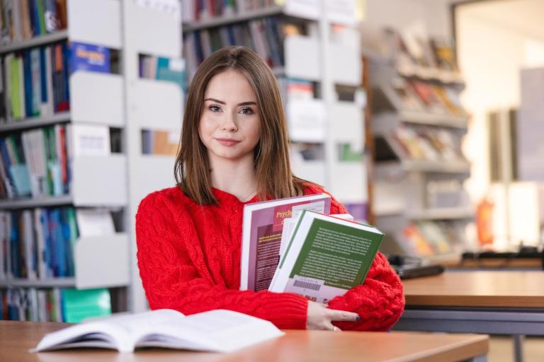Maturzyści | Uniwersytet WSB Merito Szczecin: Studentka Uniwersytetu WSB Merito trzyma książki siedząc w bibliotece.