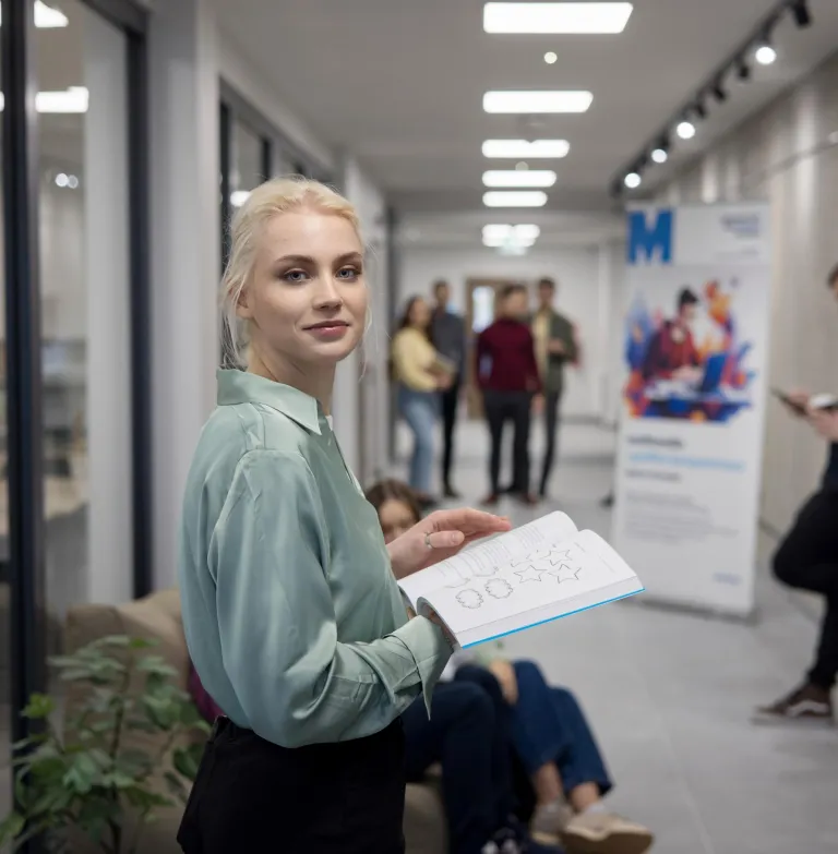 Szkoły | Uniwersytet WSB Merito Szczecin: Studentka Uniwersytetu WSB Merito przegląda książkę na korytarzu uczelni.