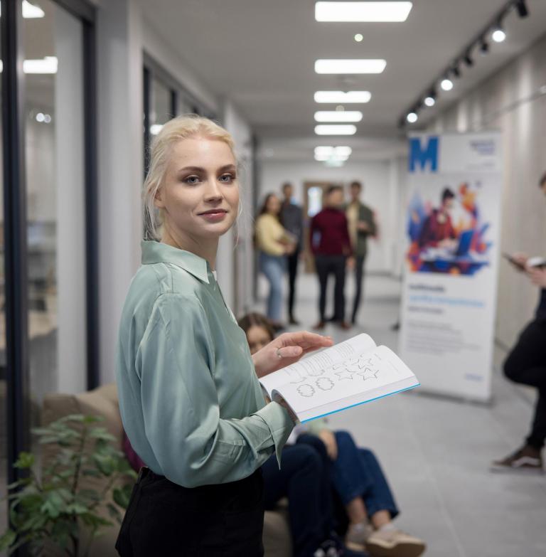 Szkoły | Uniwersytet WSB Merito Szczecin: Studentka Uniwersytetu WSB Merito przegląda książkę na korytarzu uczelni.