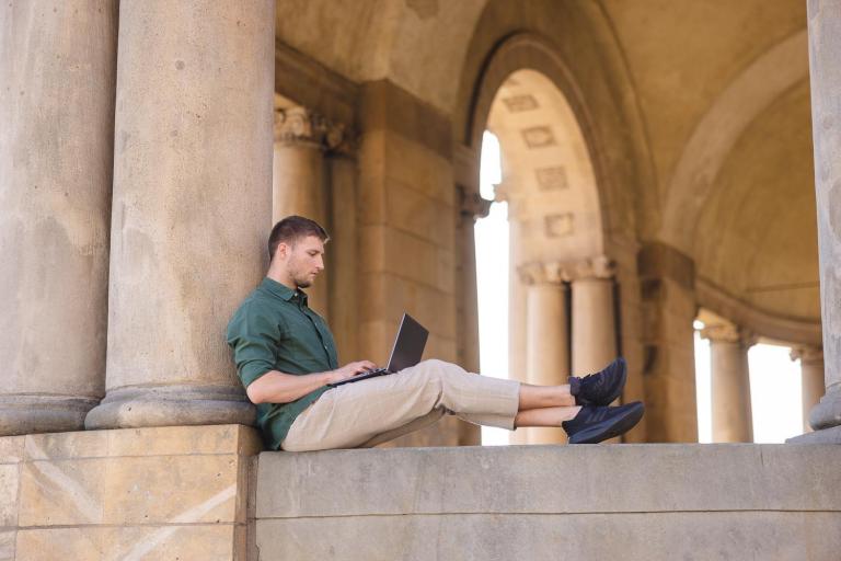 Pośrednictwo pracy | Uniwersytet WSB Merito Szczecin: Mężczyzna siedzi na murku z laptopem, pracując w otoczeniu zabytkowych kolumn.
