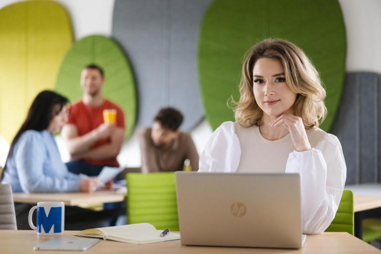 Zarządzanie i obrót nieruchomościami | Studia I stopnia w Szczecinie | Uniwersytet WSB Merito Szczecin: Studentka pracuje przy laptopie w nowoczesnej przestrzeni z innymi osobami w tle.