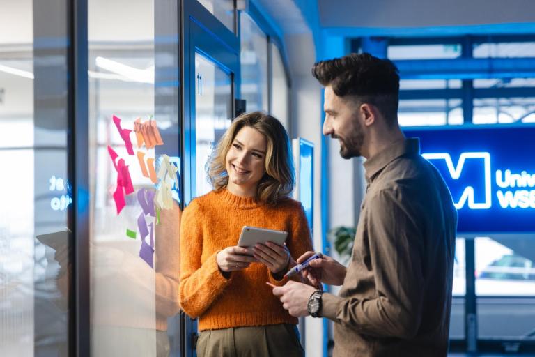 Zniżki i dofinansowania | Uniwersytet WSB Merito Szczecin: Dwoje studentów omawia notatki na szybie z kolorowymi karteczkami w nowoczesnym wnętrzu uczelni.