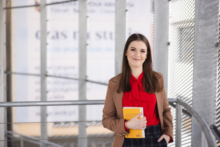 Stypendia i zniżki | Uniwersytet WSB Merito Szczecin: Uśmiechnięta studentka w budynku uczelni trzyma książkę