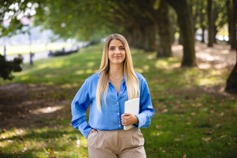 Doradca w procesie sukcesji | Studia podyplomowe w Szczecinie | Uniwersytet WSB Merito Szczecin: Grafika główna: WSB Merito Szczecin, dla nowych studentów, studentka idzie przez park trzymając książkę w ręku