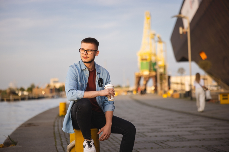 Strona główna | Uniwersytet WSB Merito Szczecin: Student WSB Merito siedzi na nadbrzeżu, w tle żurawie stoczni