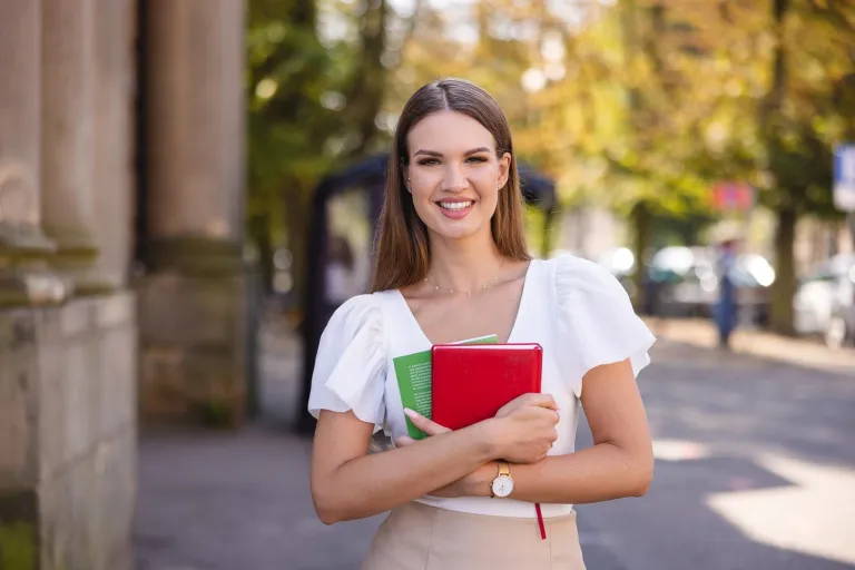 Administracja i bezpieczeństwo wewnętrzne | Uniwersytet WSB Merito Szczecin: Studentka stoi na tle miejskiej ulicy z zeszytami w rękach, uśmiechając się do obiektywu.