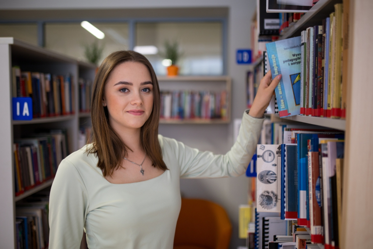ABC studenta | Uniwersytet WSB Merito w Toruniu: Grafika główna: WSB Merito Toruń, dla nowych studentów, studentka w bibliotece wybiera książkę z półki