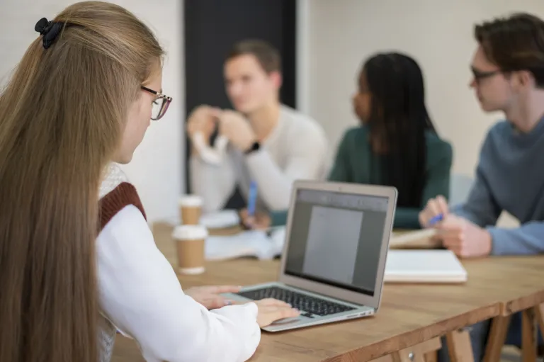 Zarządzanie personalne i płacowe, studia podyplomowe: Kobieta pracuje przy komputerze, w tle grupa osób z książkami