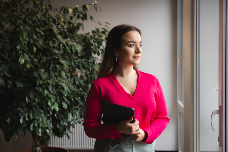 Strefa studenta | Uniwersytet WSB Merito we Wrocławiu: Studentka WSB Merito trzyma komputer w ręce i patrzy przez okno w budynku uczelni